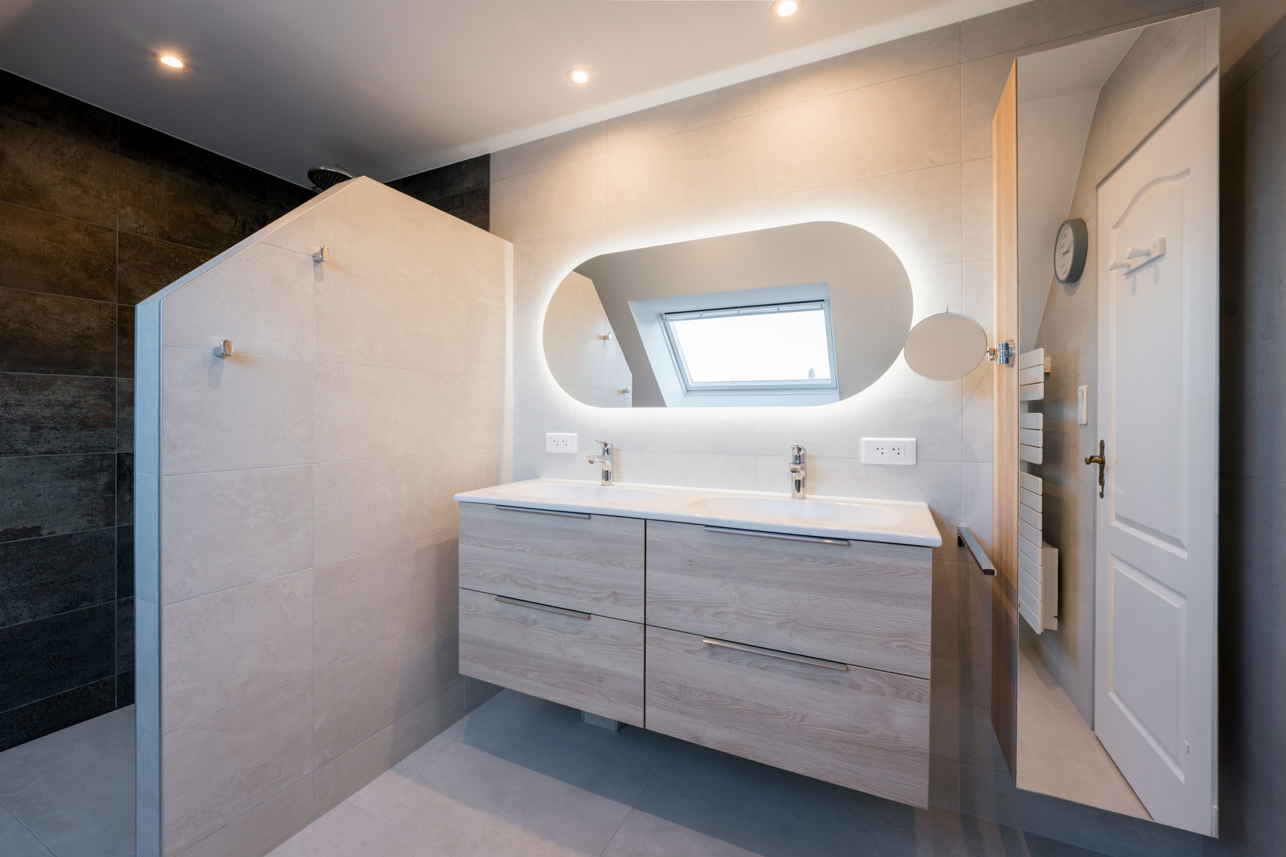 Rénovation d'une salle de bain sous pente à Mulhouse avec douche à l'italienne, meuble double vasque en bois clair et miroir rétroéclairé ovale.